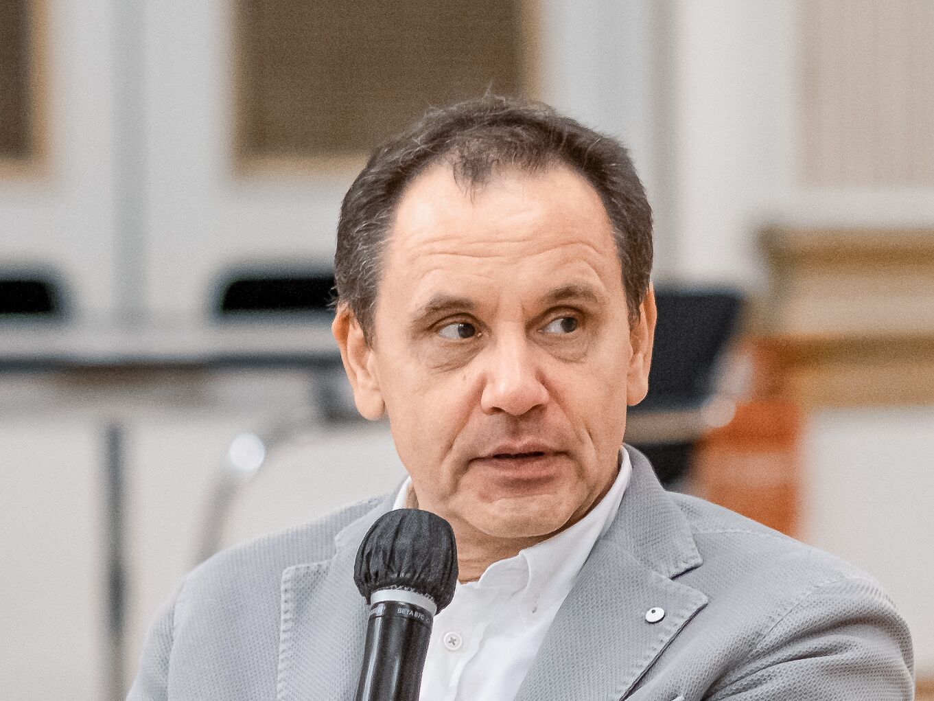 Ein Mann mittleren Alters in einem hellgrauen Sakko und weißem Hemd hält ein Mikrofon. Er schaut ernst nach rechts. Im Hintergrund sind unscharf Möbel und Teile eines Raumes sichtbar.