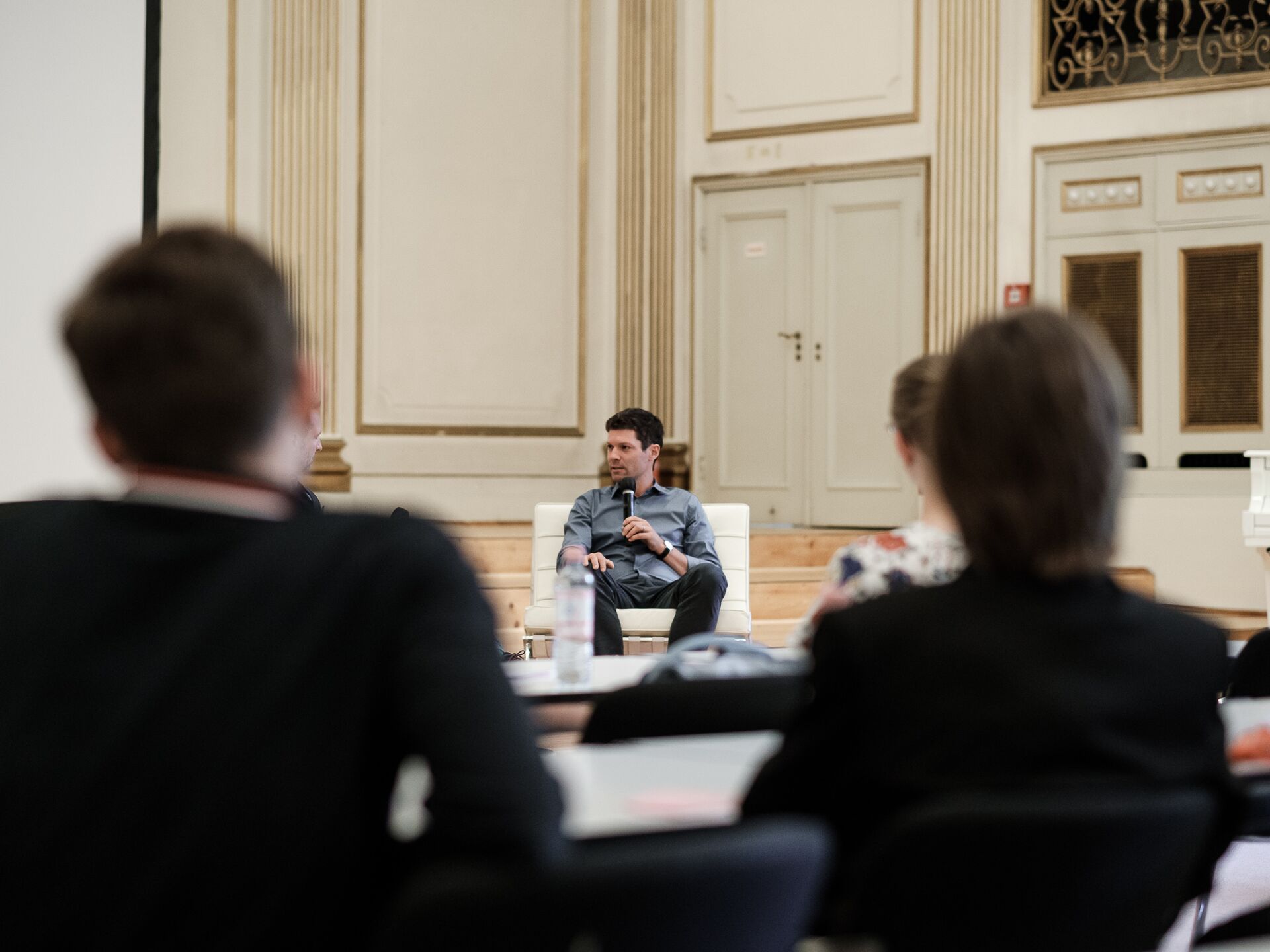 Ein Mann sitzt auf einem weißen Sessel in einem prunkvollen Saal und spricht mit einem Mikrofon. Im Vordergrund sind verschwommen mehrere Zuhörer zu sehen, die aufmerksam zuhören.