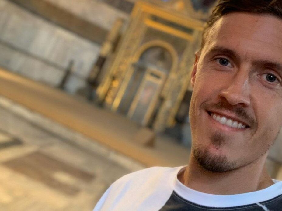 Ein junger Mann lächelt in die Kamera, im Hintergrund ist ein prachtvolles, golden verziertes Gebäude unscharf zu sehen.