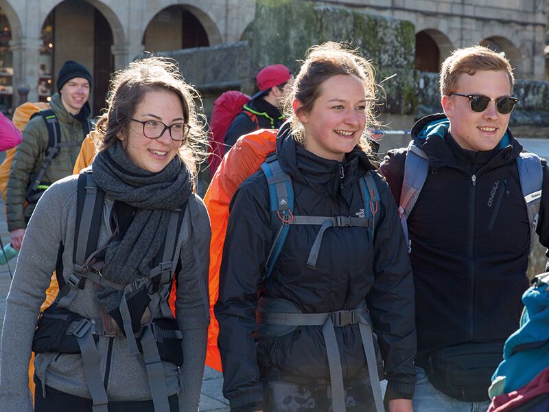 Drei junge Menschen mit Rucksäcken und Outdoor-Kleidung stehen nebeneinander und lächeln; im Hintergrund sind weitere Wanderer sowie ein steinernes Gebäude mit Arkaden zu sehen.