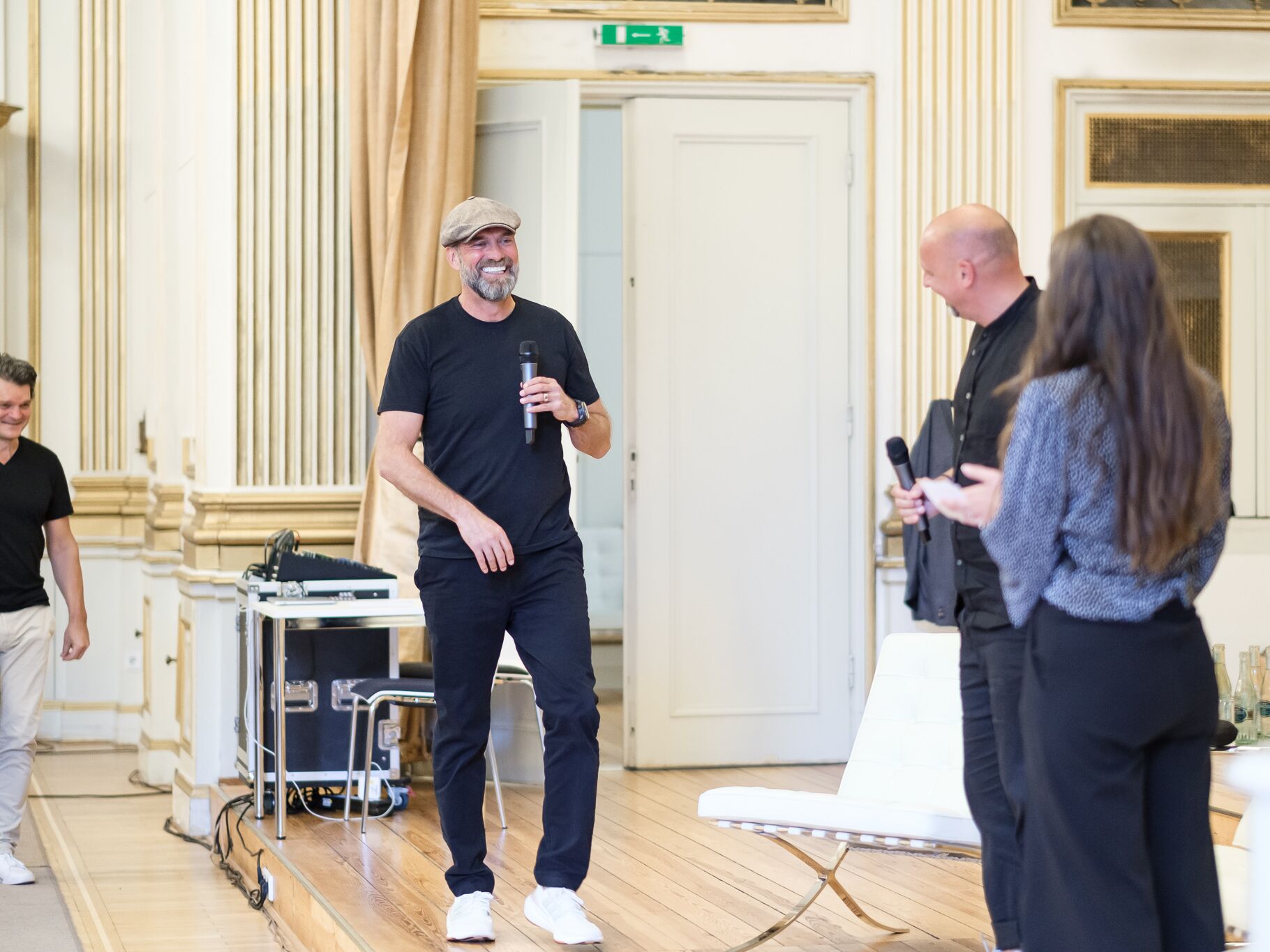 Ein Mann mit Mütze und Mikrofon betritt lächelnd eine Bühne in einem prunkvollen Saal. Zwei weitere Personen mit Mikrofonen und ein Mann im Hintergrund begrüßen ihn.