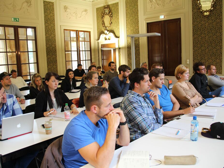 Studierende sitzen konzentriert in einem historischen Seminarraum und hören aufmerksam einer Vorlesung zu.