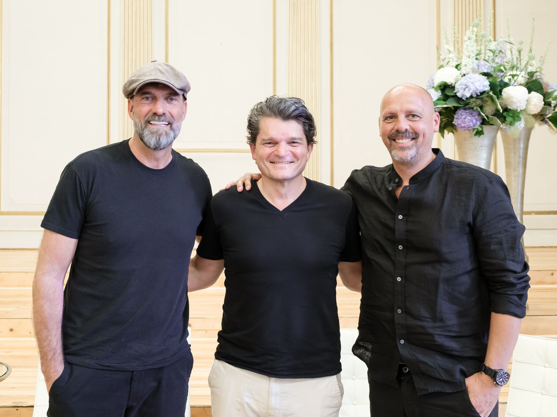 Drei Männer stehen nebeneinander und lächeln in die Kamera. Der Mann links trägt eine Schirmmütze und ein schwarzes T-Shirt, der Mann in der Mitte ein schwarzes T-Shirt mit heller Hose, und der Mann rechts ein schwarzes Hemd. Im Hintergrund sind große Blu