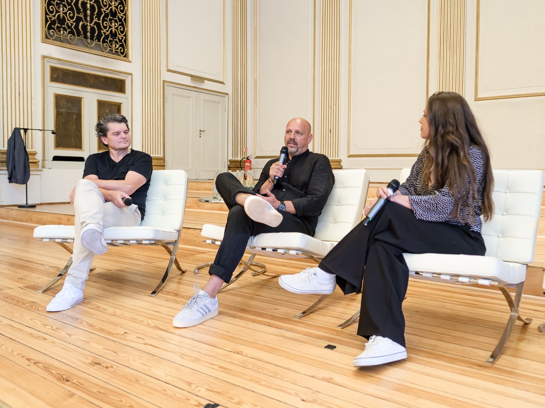Drei Personen sitzen mit Mikrofonen in Sesseln auf einer Bühne und führen ein Gespräch in einem Saal.
