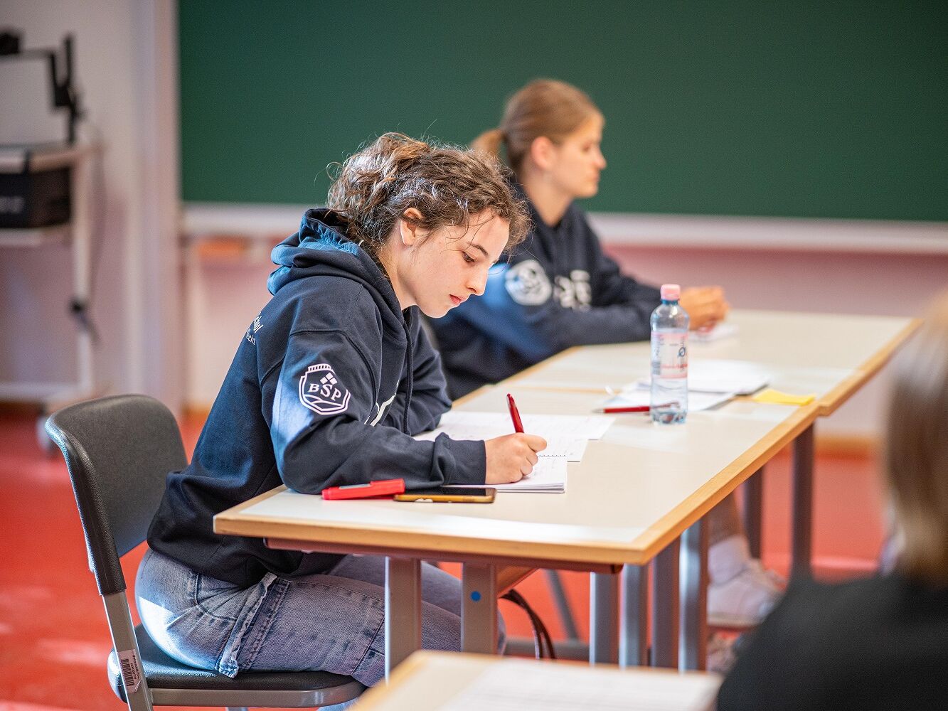 Eine junge Frau in einem BSP-Hoodie sitzt in einem Seminarraum an einem Tisch, schreibt konzentriert in ein Heft, daneben liegen Textmarker. Im Hintergrund sitzt eine weitere Person, ebenfalls im Hoodie, vor einer Tafel.