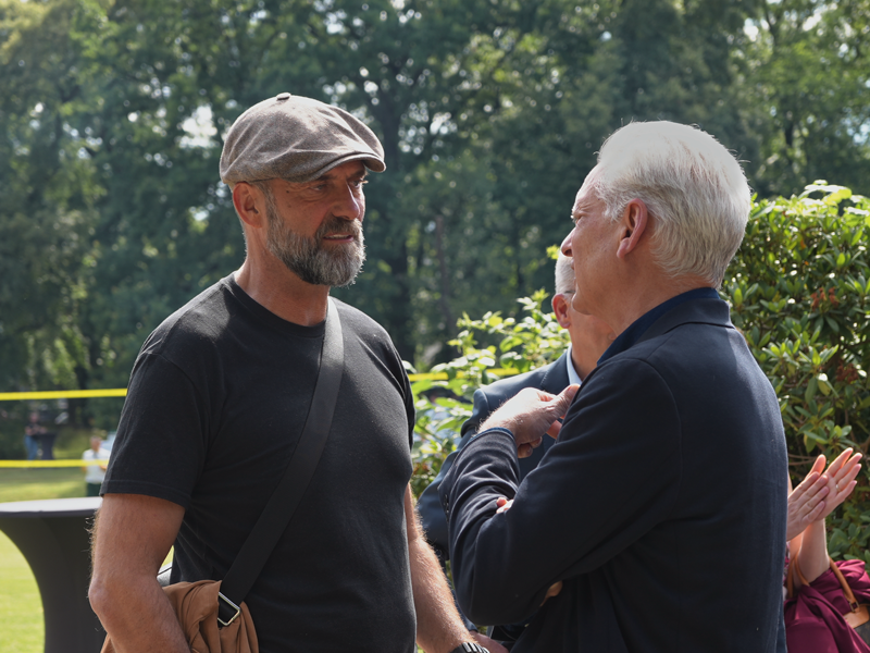 Zwei Männer stehen im Freien und unterhalten sich. Einer trägt ein schwarzes T-Shirt und eine Schirmmütze, der andere einen dunkelblauen Blazer. Im Hintergrund sind Bäume und Sträucher zu sehen.