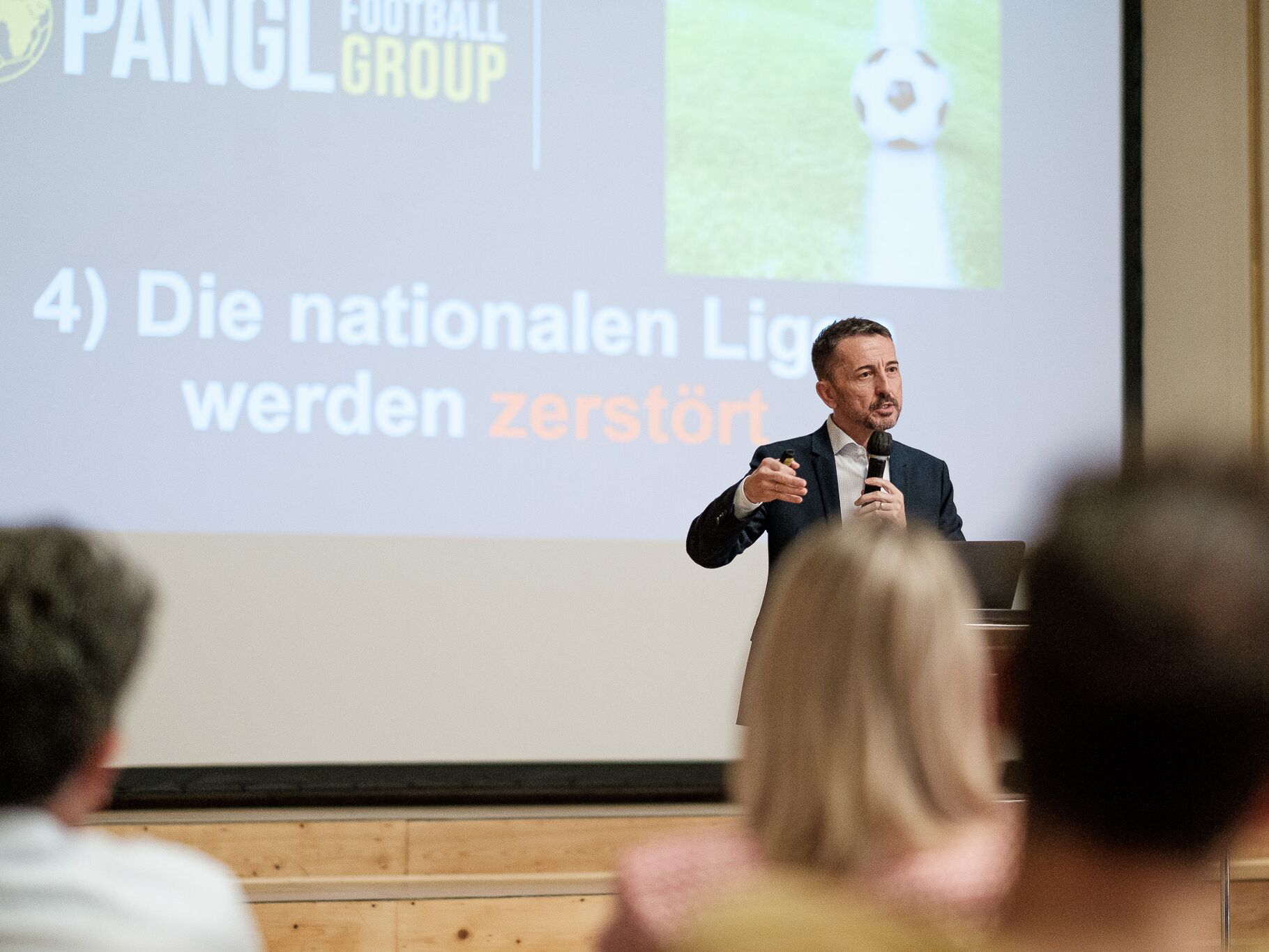 Ein Mann im Anzug hält einen Vortrag mit Mikrofon, hinter ihm eine Präsentation mit der Aufschrift „Die nationalen Ligen werden zerstört“.