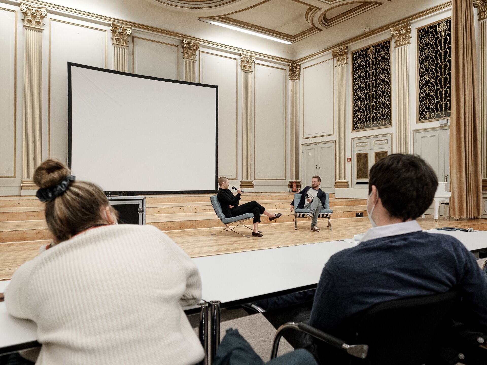 Eine Podiumsdiskussion in einem prunkvollen Saal. Zwei Personen sitzen vorne auf Sesseln mit Mikrofonen, während das Publikum auf Stühlen an Tischen zuhört. Im Hintergrund ist eine große, leere Leinwand sichtbar, die Bühne ist mit Holzstufen gestaltet.