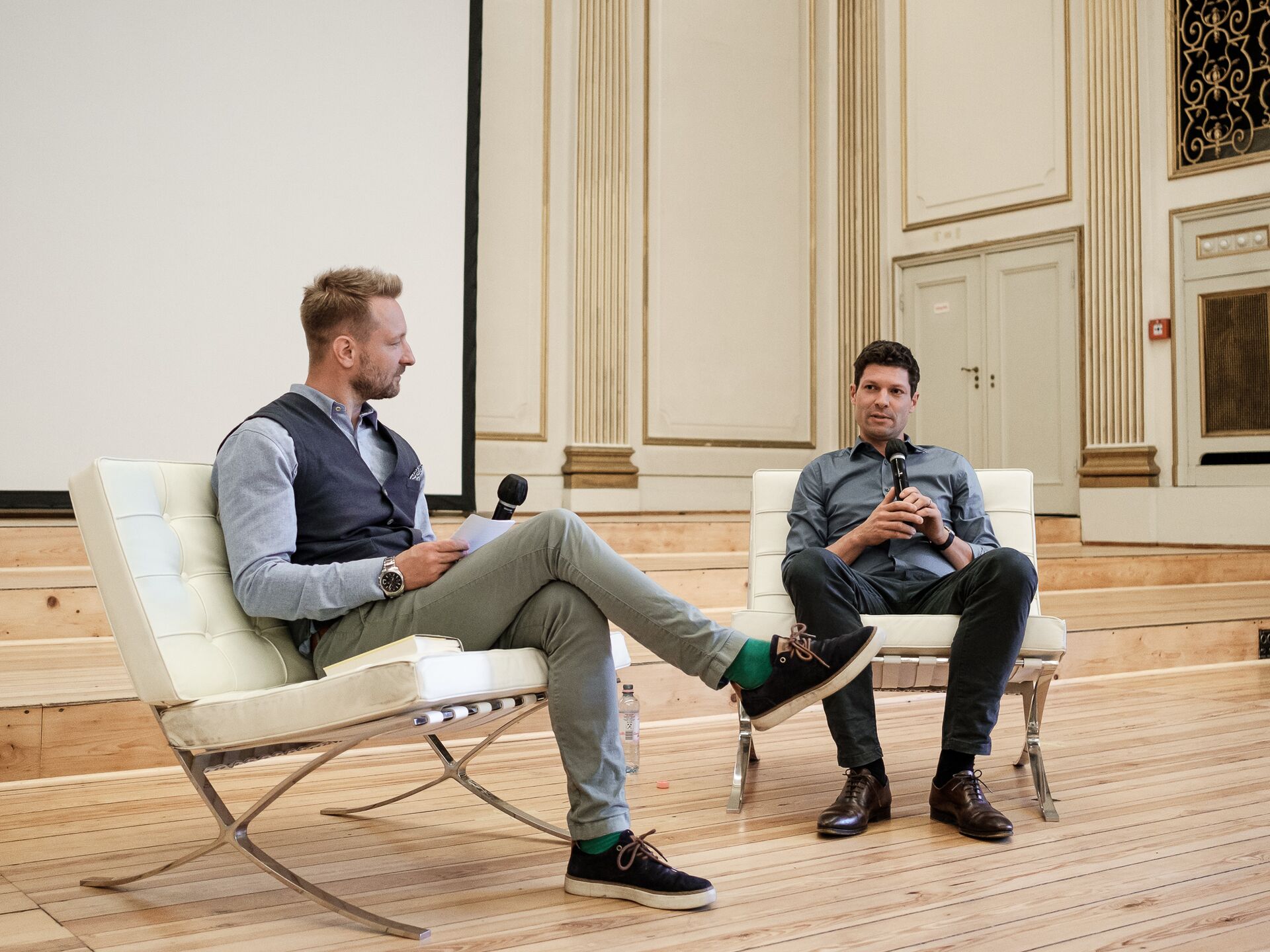 Zwei Männer sitzen in weißen Sesseln auf einer Bühne und führen ein Gespräch mit Mikrofonen. Der Mann links trägt ein hellblaues Hemd, eine graue Weste, grüne Hose und schwarze Sneaker mit grünen Socken. Der Mann rechts trägt ein dunkelblaues Hemd, schwar