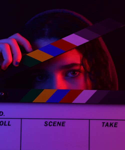 Eine Person blickt durch eine bunte Filmklappe, die ihr Gesicht teilweise verdeckt, mit dramatischer Beleuchtung in Rot und Blau.