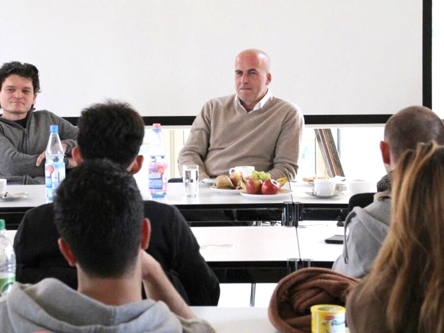Zwei Männer sitzen an einem Tisch vor einer Projektionsfläche, während mehrere Personen ihnen zuhören. Auf dem Tisch stehen Wasserflaschen, Obst und Gebäck.
