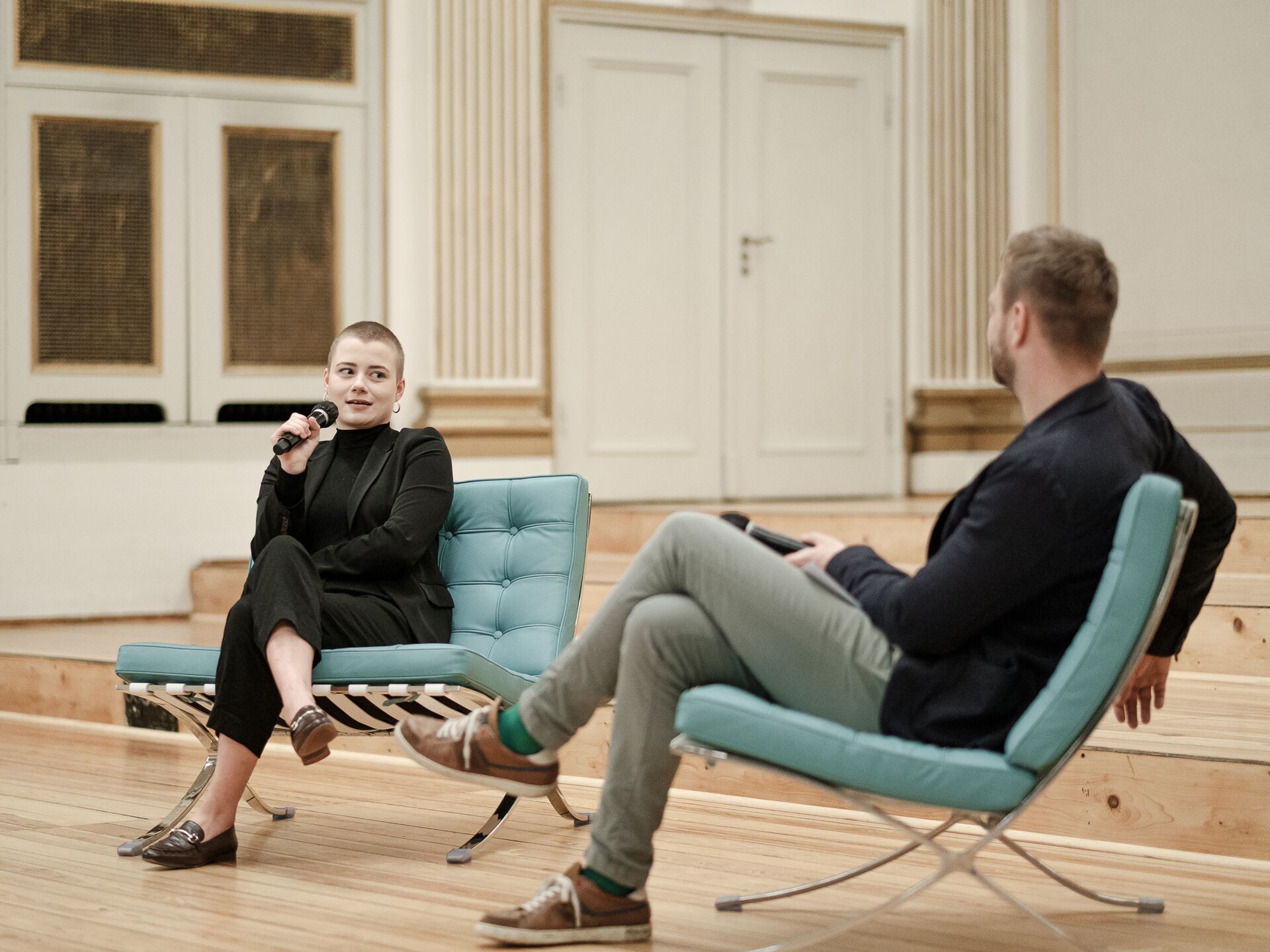Zwei Personen sitzen auf modernen Sesseln in einem großen Saal und führen ein Gespräch. Die Person links mit kurz geschorenen Haaren trägt einen schwarzen Anzug und spricht in ein Mikrofon. Die Person rechts hört aufmerksam zu und hält ebenfalls ein Mikro