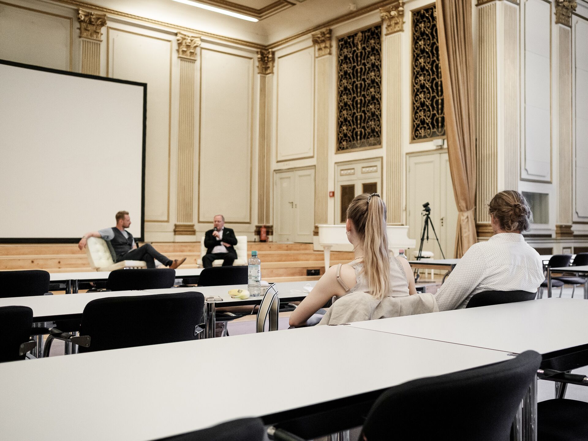 Zwei Männer sitzen auf einer Bühne in einem prunkvollen Saal und führen ein Gespräch mit Mikrofonen. Im Vordergrund sitzen einige Zuhörer an Tischen, während eine Kamera auf der rechten Seite aufgestellt ist.