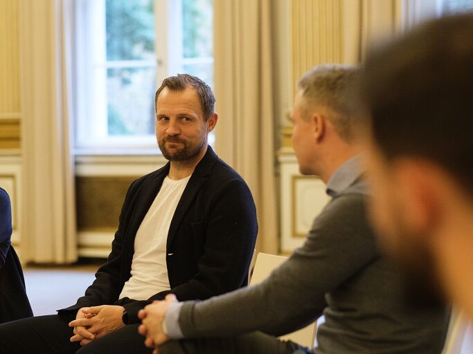 Männer sitzen in einem Stuhlkreis und führen ein Gespräch in einem hellen Raum mit großen Fenstern.