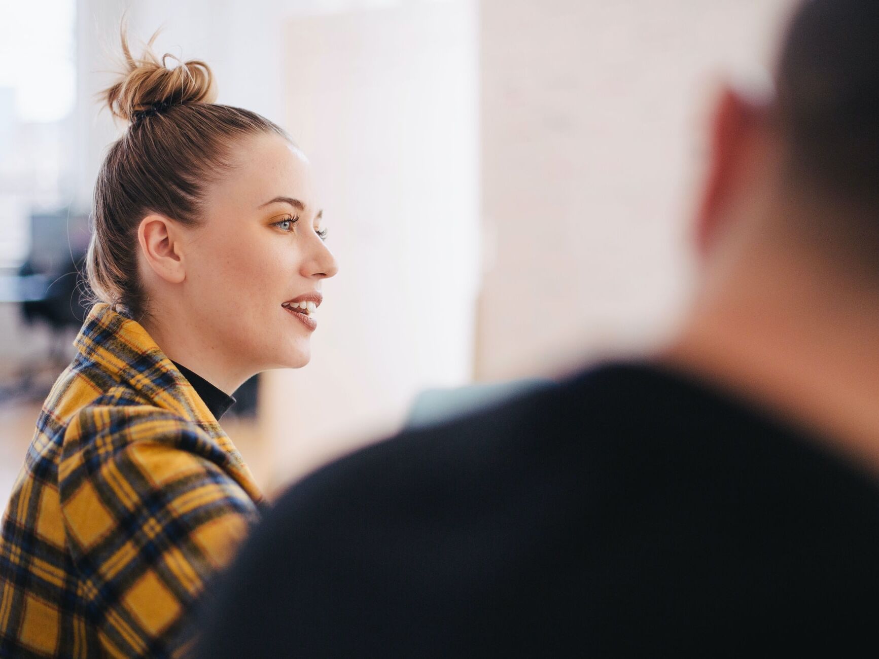 Junge Frau mit Dutt und gelb-blau karierter Jacke spricht, im Vordergrund ist unscharf der Hinterkopf einer weiteren Person zu sehen.