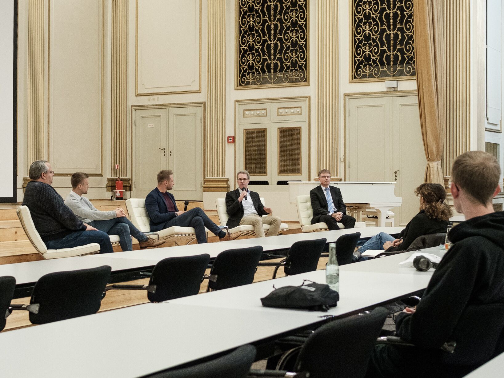 Fünf Männer sitzen bei einer Podiumsdiskussion auf einer Bühne, vor ihnen hört ein kleines Publikum zu.