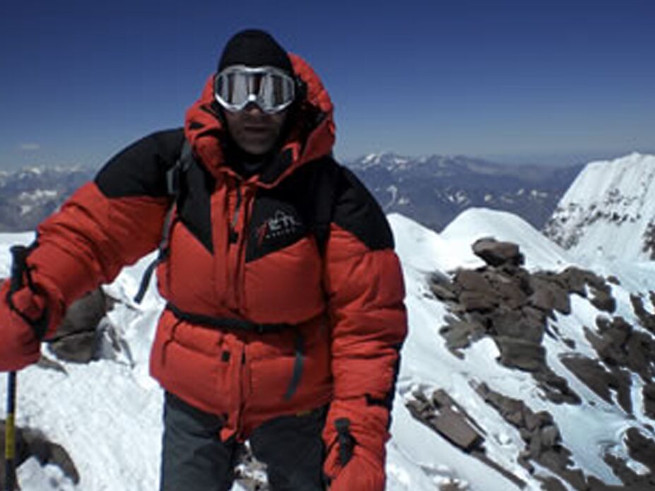 Ein Bergsteiger in dicker roter und schwarzer Expeditionsjacke mit Skibrille steht auf einem verschneiten Berggipfel, umgeben von schneebedeckten Gipfeln und klarem blauen Himmel.