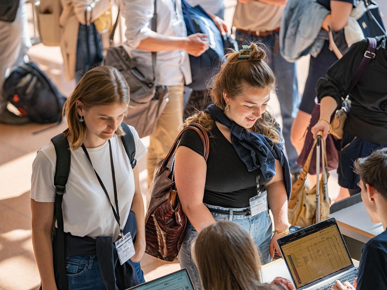 Zwei junge Frauen mit Namensschildern und Rucksäcken stehen an einem Anmeldetisch, während Personen mit Laptops ihre Daten erfassen; im Hintergrund weitere Teilnehmende mit Taschen.