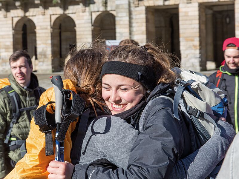 Zwei junge Wanderinnen mit großen Rucksäcken umarmen sich lächelnd vor einem historischen Gebäude, während andere Menschen im Hintergrund stehen.