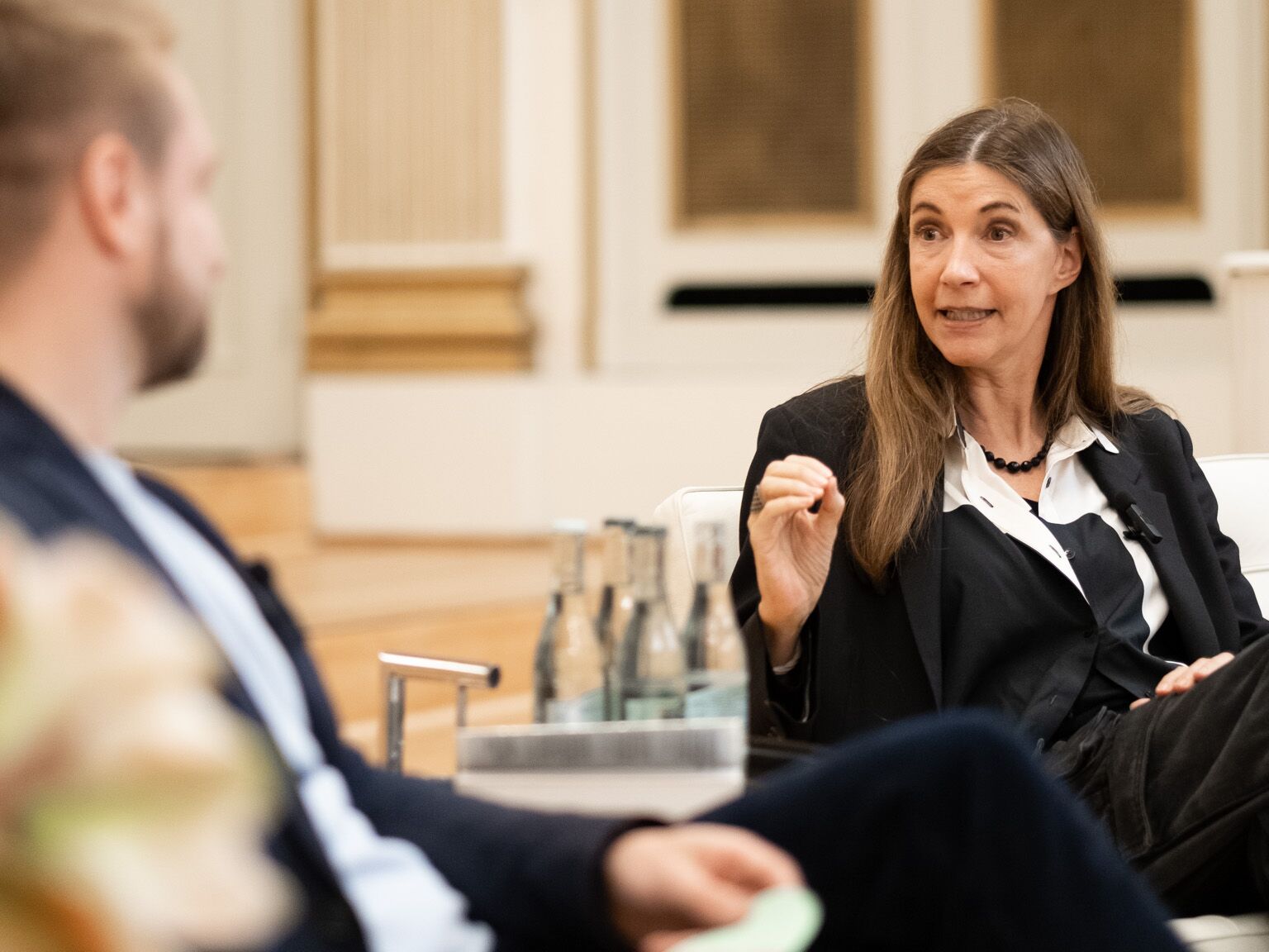 Frau im Anzug spricht gestikulierend in einer Podiumsdiskussion, neben ihr sitzt ein weiterer Teilnehmer.
