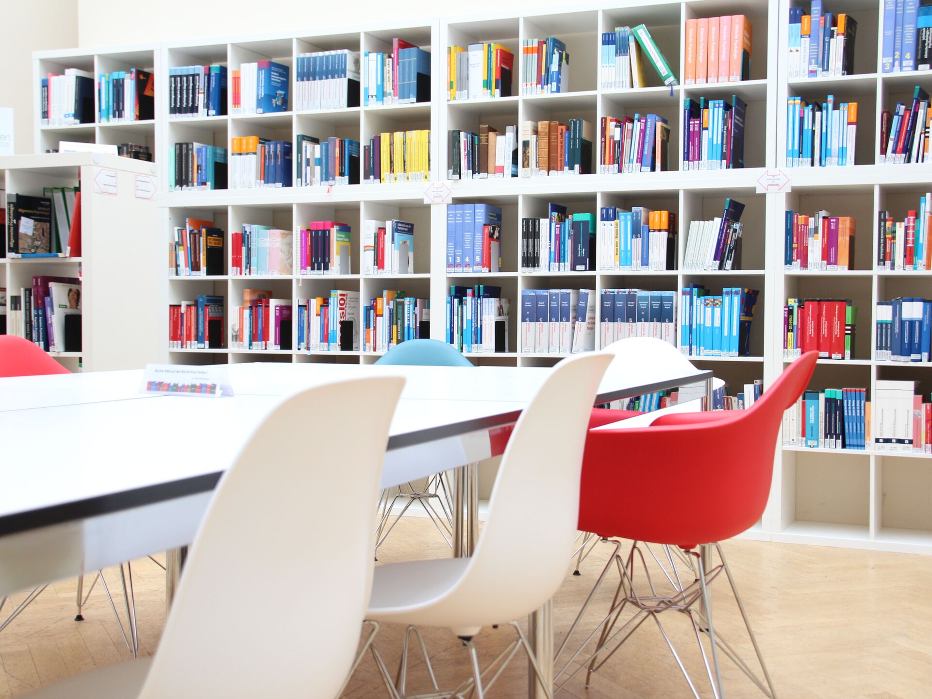 Helle Bibliothek mit Bücherregalen, weißen Tischen und bunten Stühlen.