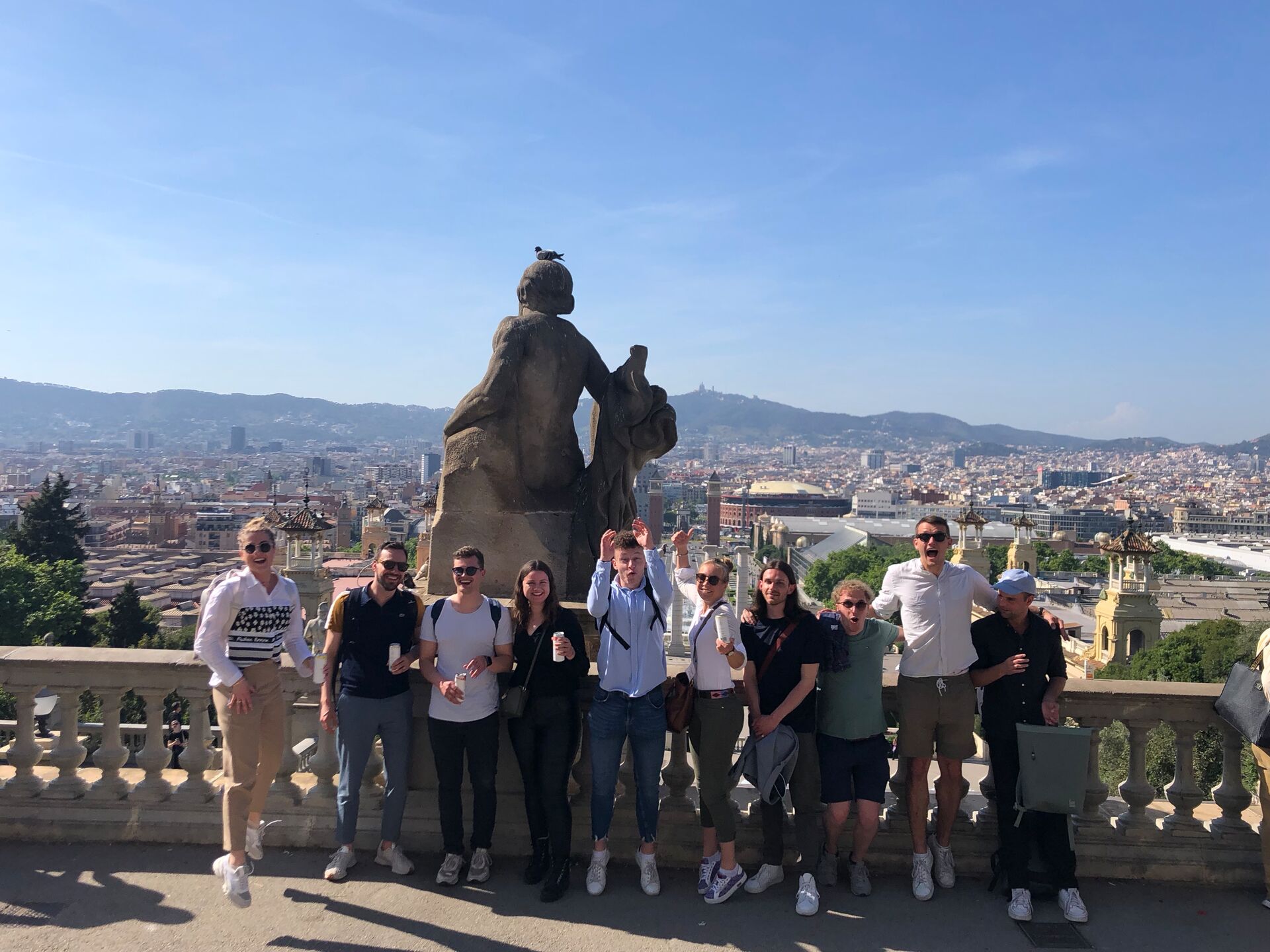 Eine Gruppe junger Menschen posiert lachend vor einer Statue mit Blick auf die Stadt Barcelona.