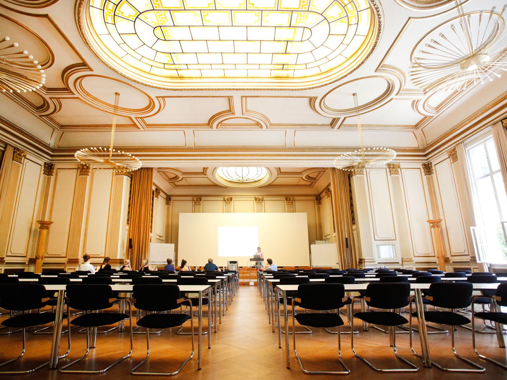 Ein großer, prunkvoller Saal mit hohen Decken, goldenen Verzierungen und einem großen verzierten Glasdach. Mehrere Reihen schwarzer Stühle mit weißen Tischen sind aufgestellt, vorne steht ein Redner am Pult vor einer großen Leinwand.