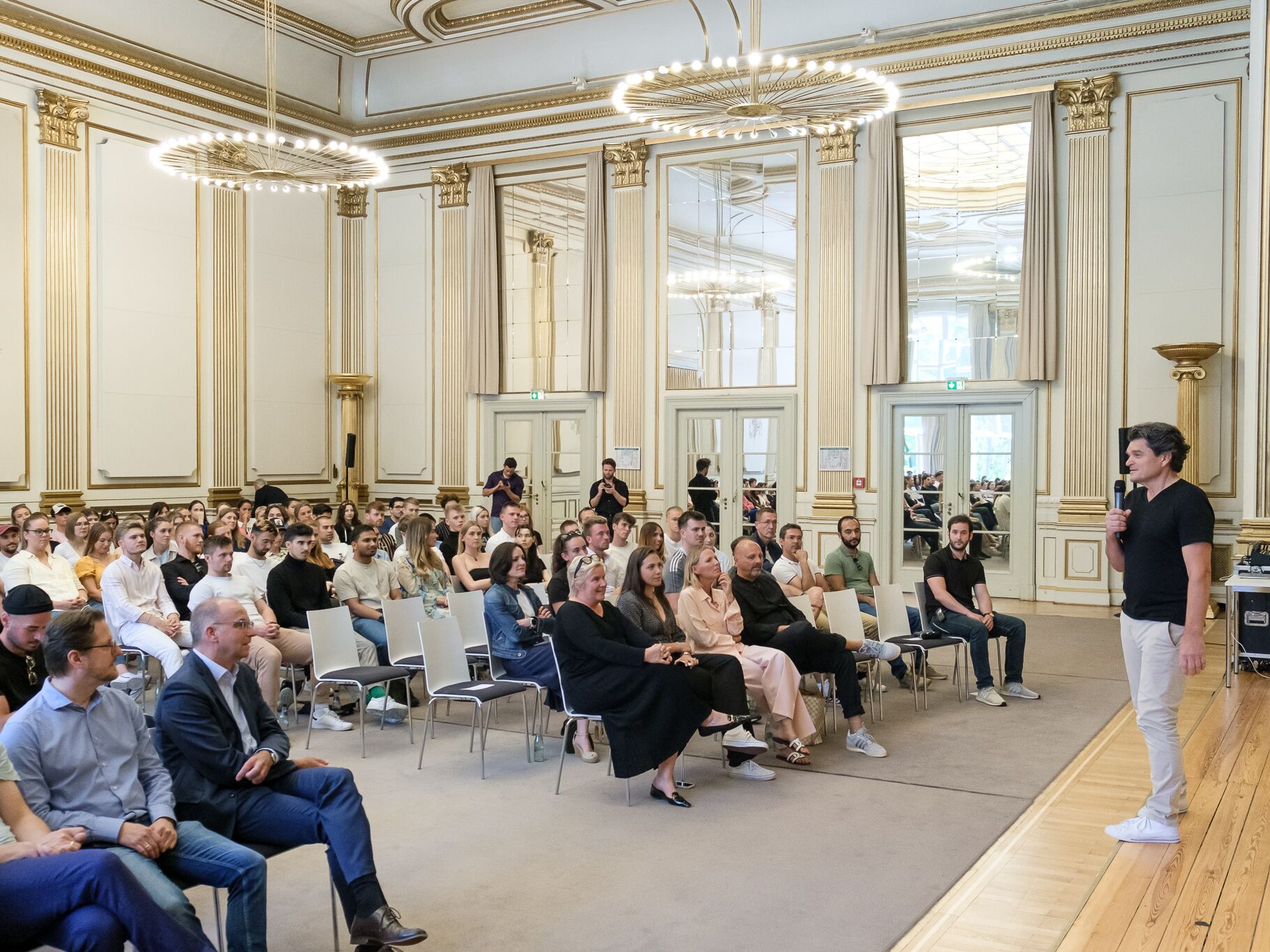 Ein Mann spricht mit einem Mikrofon vor einem großen Publikum in einem prunkvollen Saal mit goldverzierten Wänden und großen Spiegeln. Die Zuhörer sitzen aufmerksam in Reihen und hören zu.