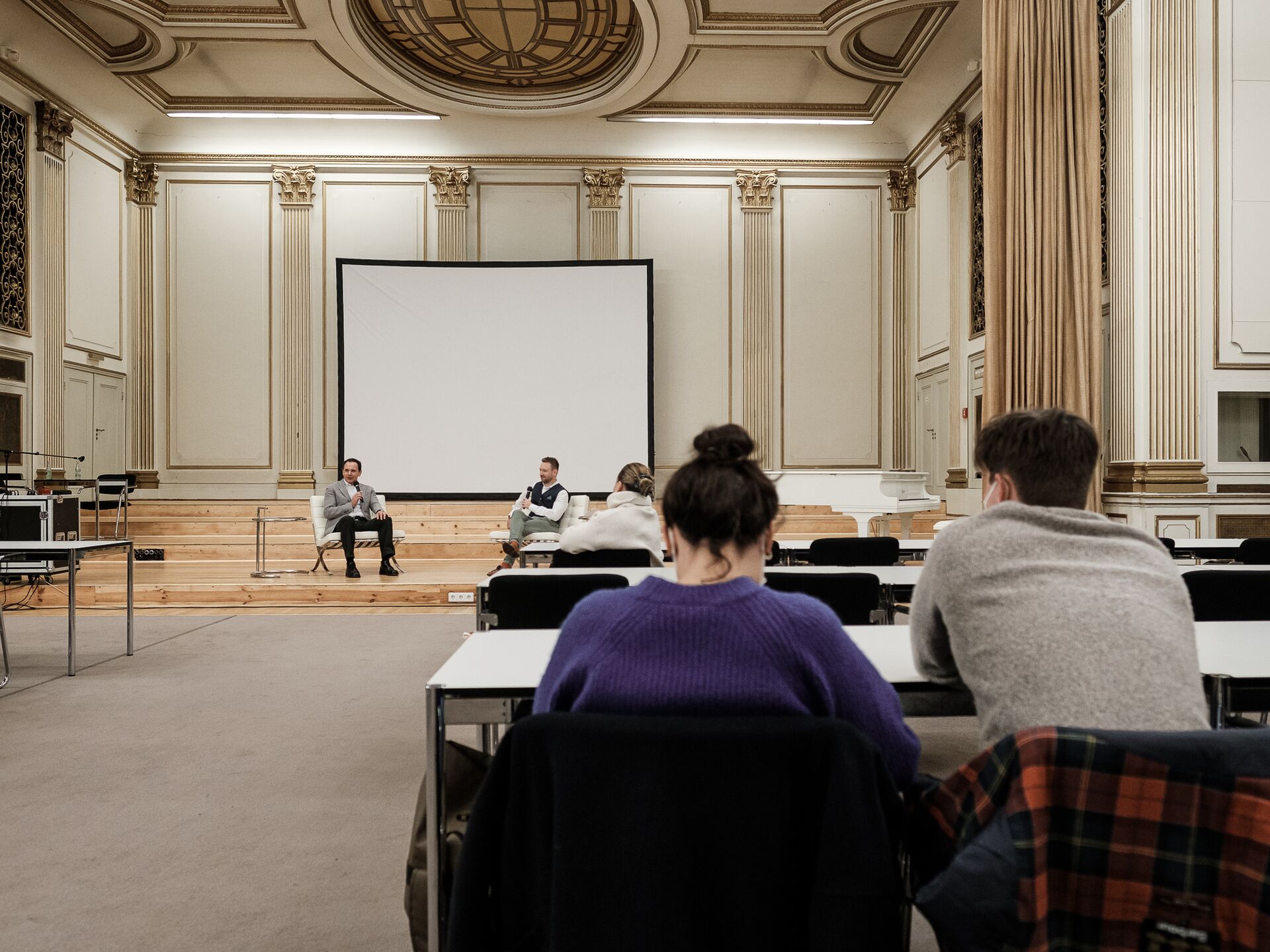 In einem großen, prunkvoll gestalteten Saal mit hohen Decken und goldenen Verzierungen sitzen zwei Männer auf einer Bühne in weißen Sesseln und sprechen mit Mikrofonen. Hinter ihnen steht eine große weiße Leinwand. Im Vordergrund sind einige Zuhörer von h