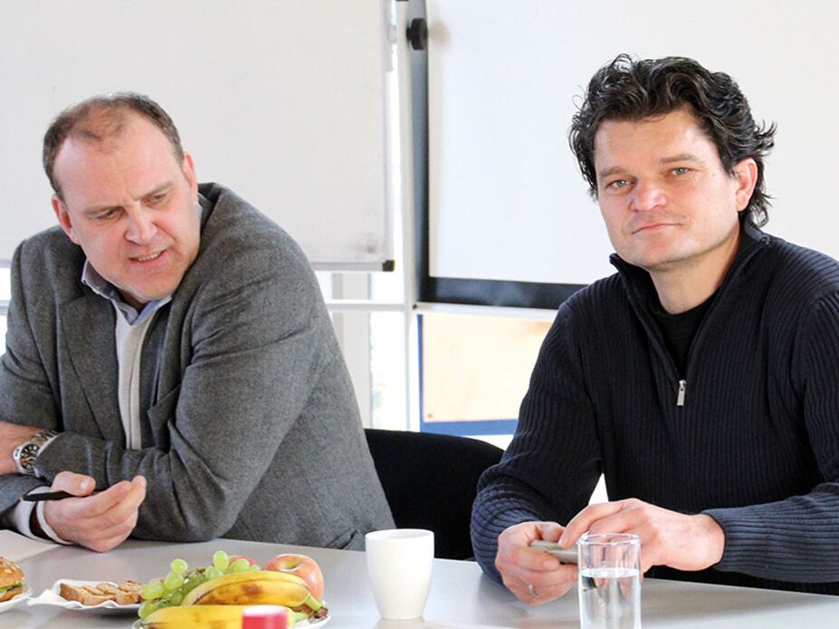 Zwei Männer sitzen an einem Tisch mit Obst, Getränken und Snacks und führen ein Gespräch in einer Besprechungsumgebung.