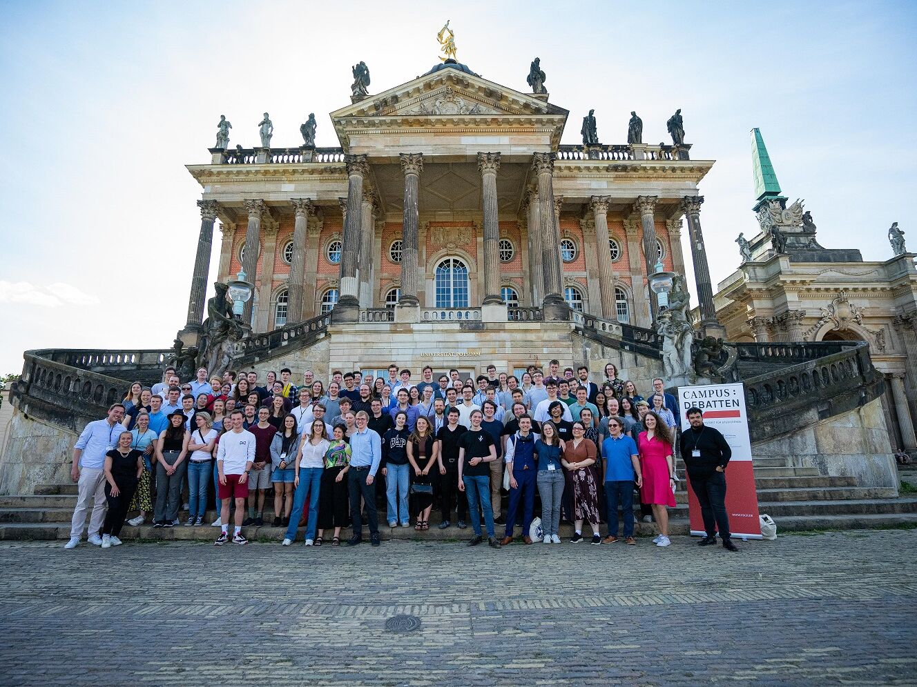 Großes Gruppenfoto von jungen Erwachsenen vor einem historischen Gebäude mit Säulen und Statuen, rechts ein Banner mit der Aufschrift "Campus: Debatten".