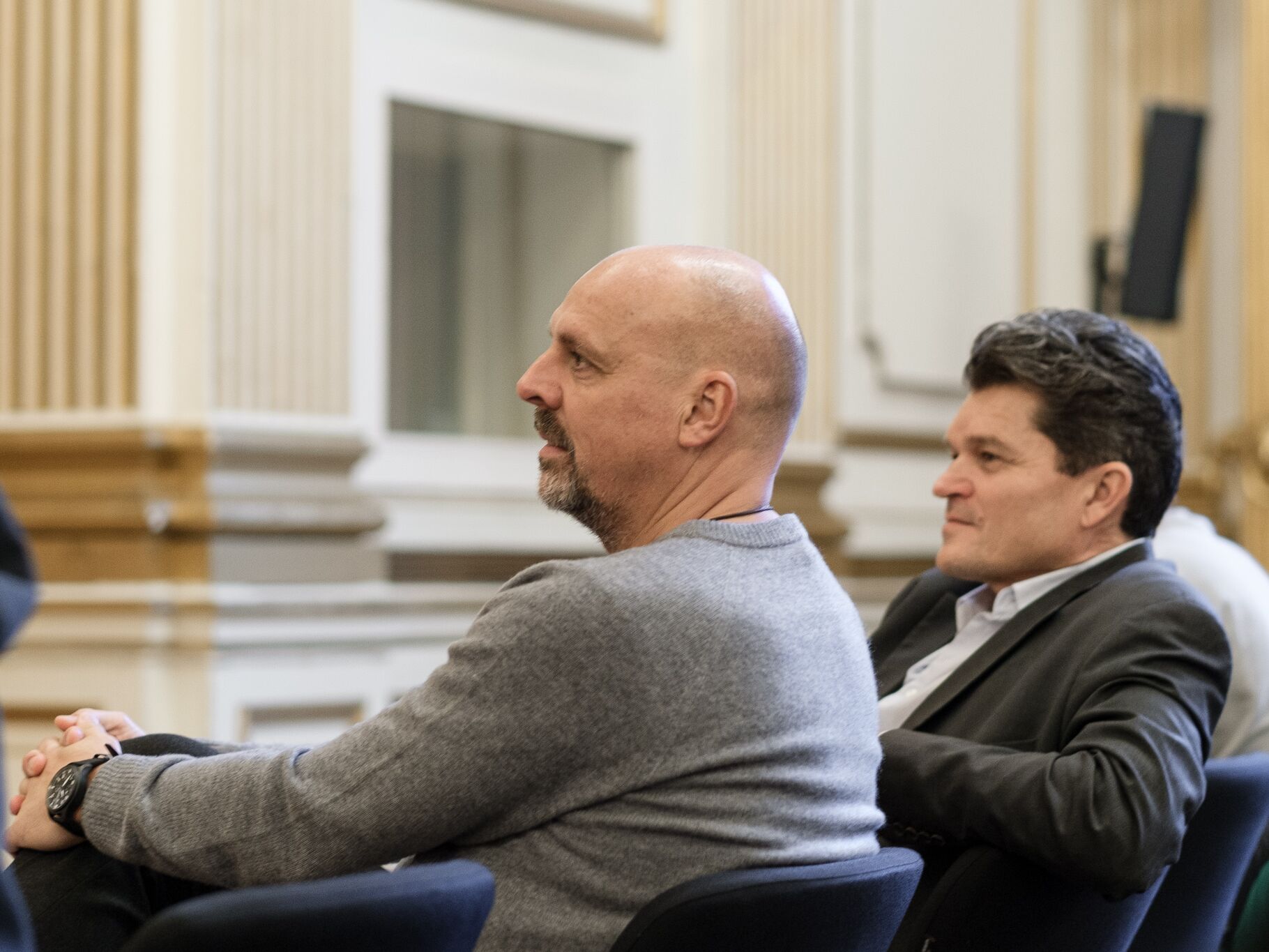 Zwei Männer sitzen nebeneinander in einem festlichen Saal und hören aufmerksam einer Veranstaltung oder Diskussion zu.
