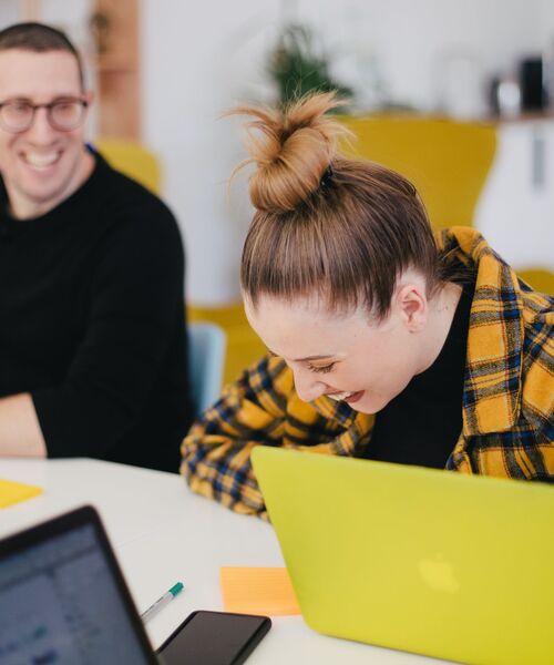 Zwei Personen sitzen lachend an einem Tisch mit Laptops und Notizen, in einer freundlichen Arbeitsumgebung.
