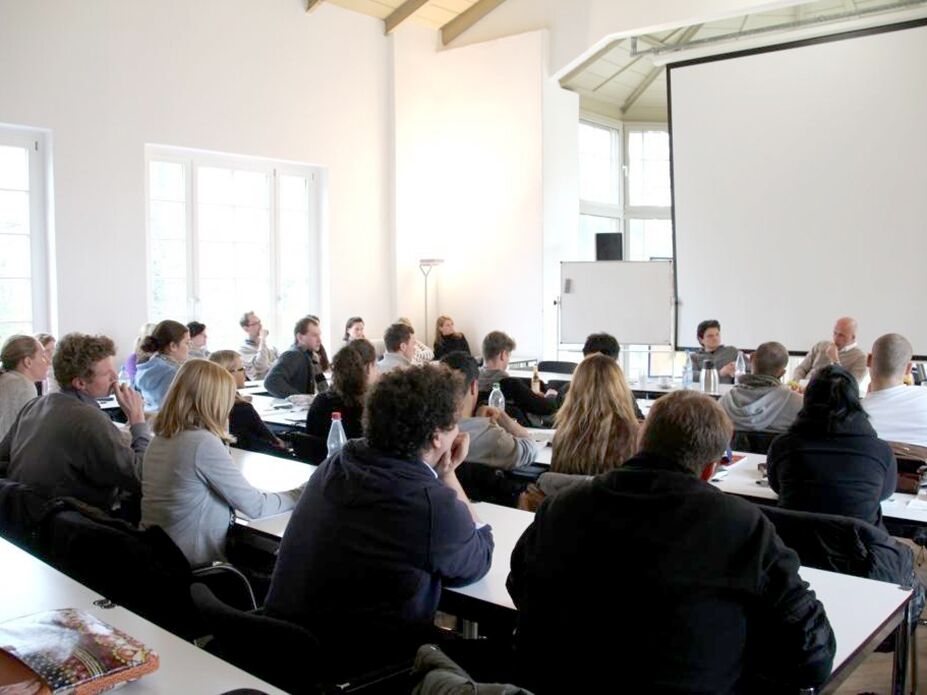 Studierende folgen aufmerksam einer Vorlesung in einem hellen Seminarraum mit großer Leinwand.