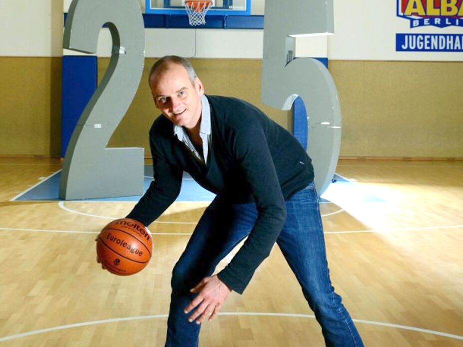 Ein Mann steht auf einem Basketballfeld, hält einen Basketball in der Hand und posiert spielerisch, im Hintergrund große Zahlen und ein Basketballkorb.