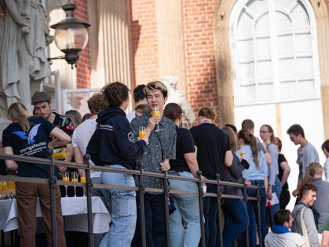 Eine Gruppe junger Menschen steht draußen vor einem historischen Gebäude bei einem Empfang. Einige halten Gläser mit Orangensaft in der Hand, im Vordergrund unterhalten sich zwei Personen. Links wird am Tisch weiterer Saft eingeschenkt.