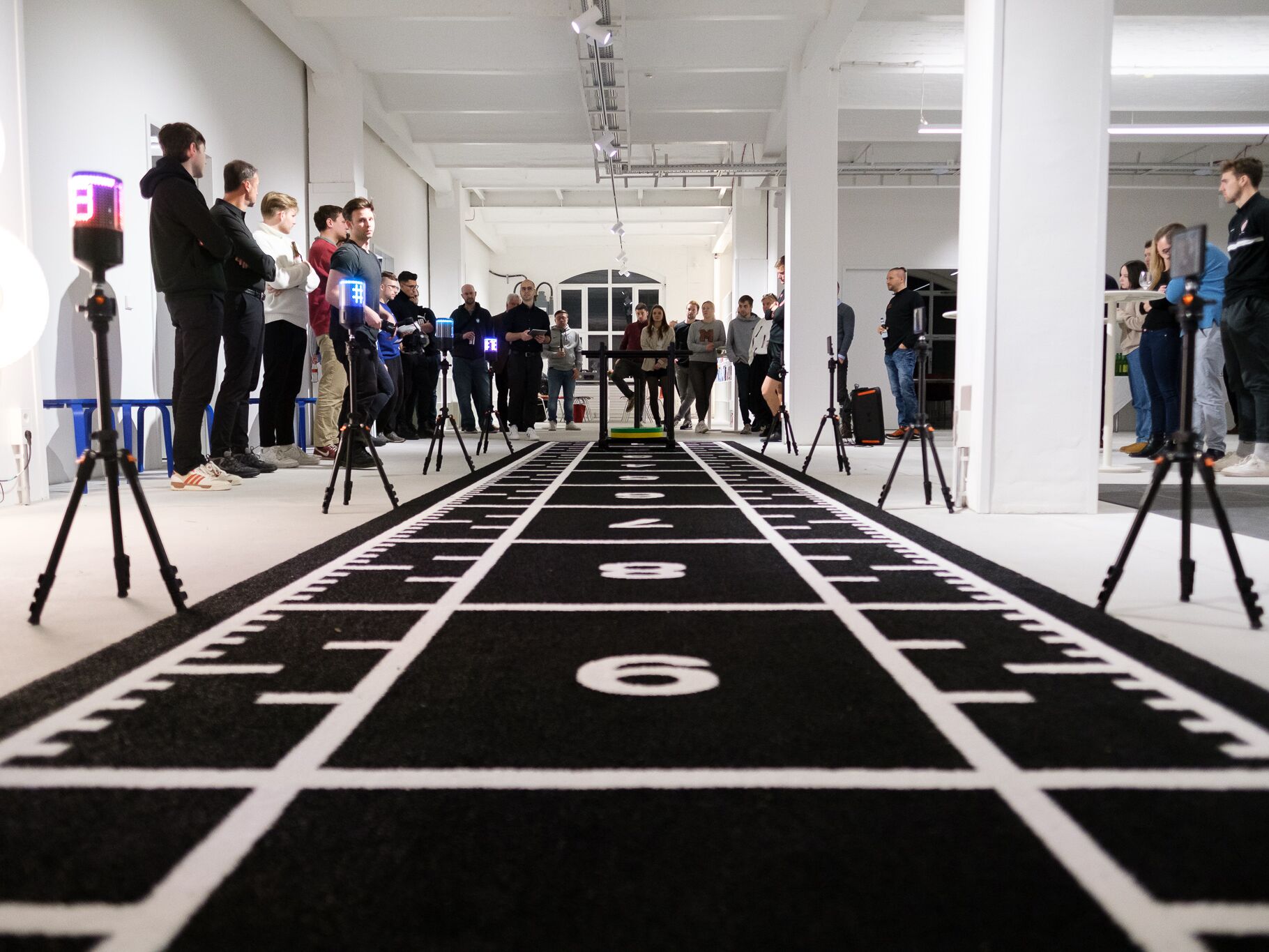Menschen versammeln sich in einer Halle um eine schwarze Sprintbahn mit Messgeräten.