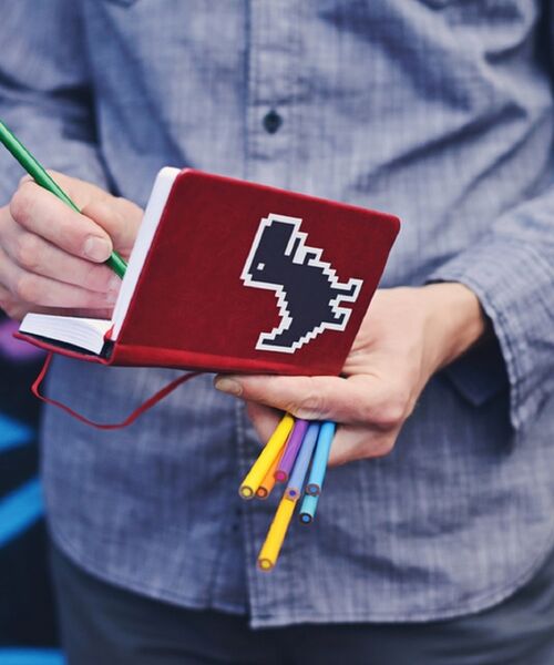 Person hält ein rotes Notizbuch mit Pixel-Art-Aufkleber eines Dinosauriers und schreibt mit einem grünen Buntstift, während sie mehrere bunte Stifte in der Hand trägt.