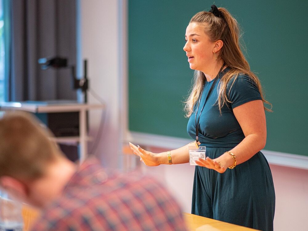 Eine junge Frau hält einen Vortrag in einem Seminarraum, spricht gestikulierend vor einer Tafel und trägt ein Namensschild um den Hals.
