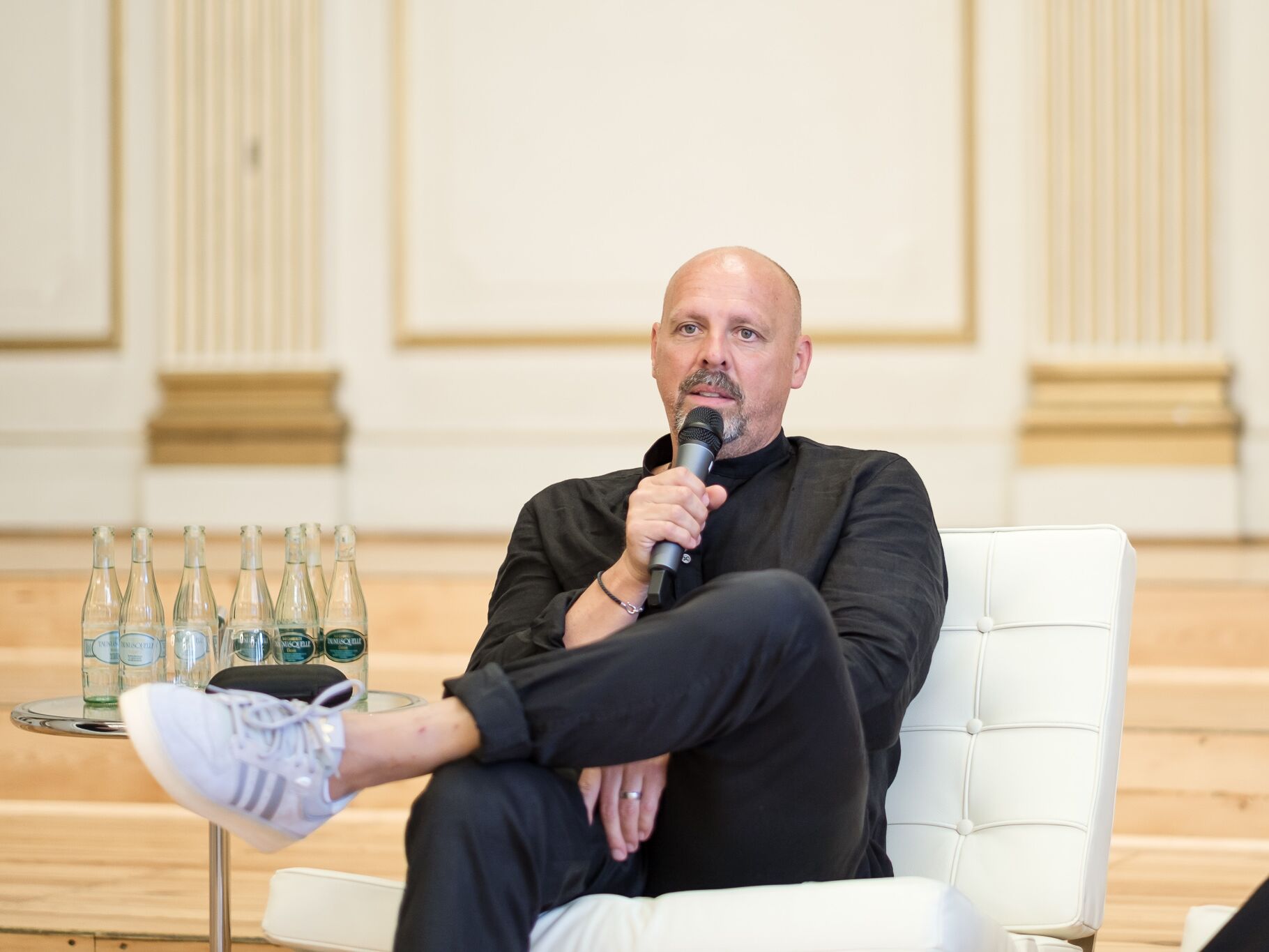 Ein Mann sitzt entspannt in einem weißen Sessel, hält ein Mikrofon in der Hand und spricht während einer Veranstaltung. Neben ihm stehen mehrere Wasserflaschen auf einem kleinen Tisch.