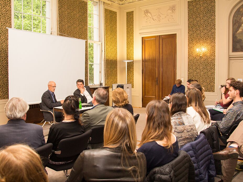 Zwei Dozenten sitzen vorne in einem prunkvollen Seminarraum neben einer großen Leinwand und diskutieren, während zahlreiche Studierende aufmerksam zuhören.