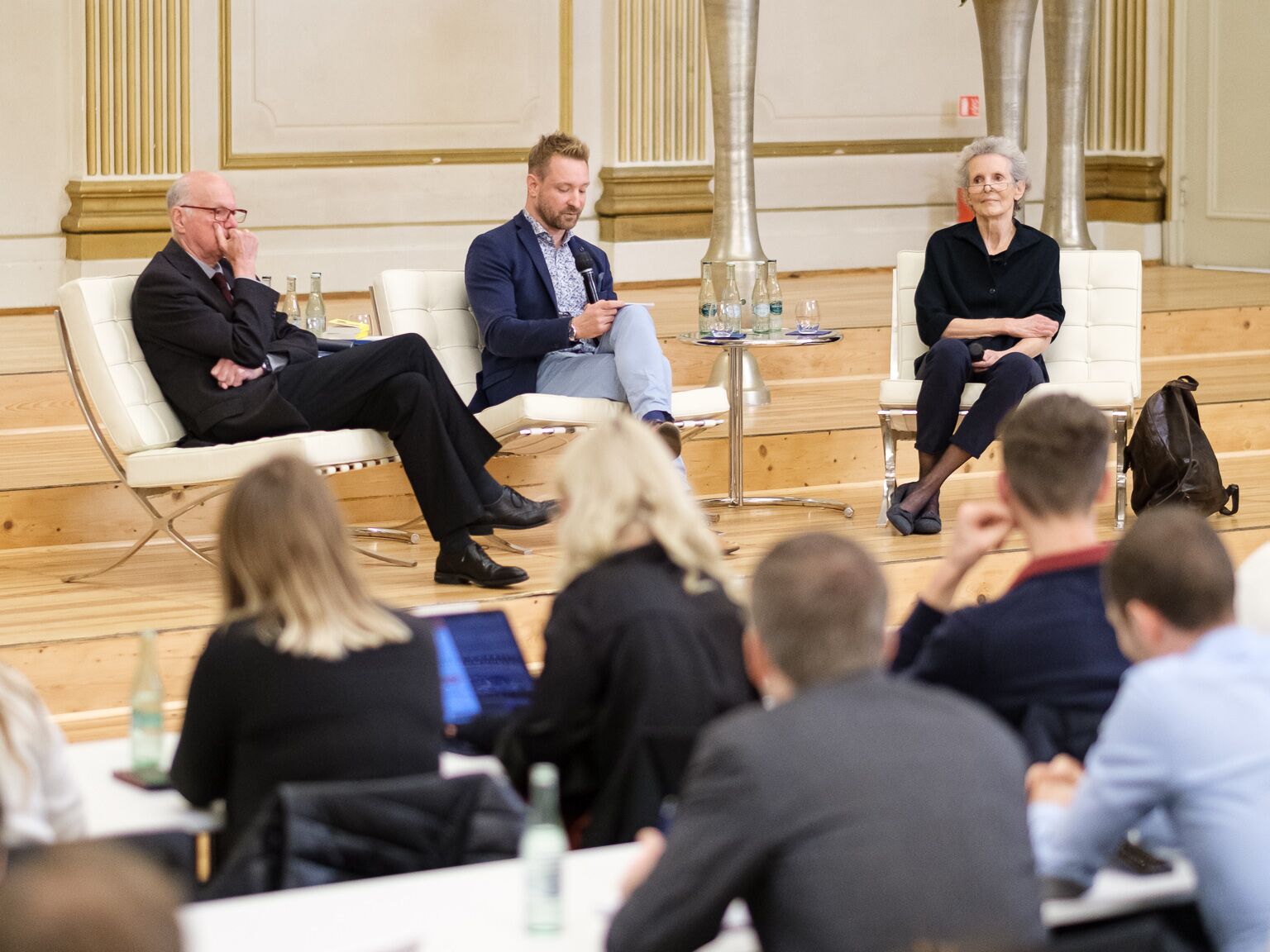 Drei Personen sitzen auf einer Bühne bei einer Podiumsdiskussion, vor ihnen hört ein Publikum zu.