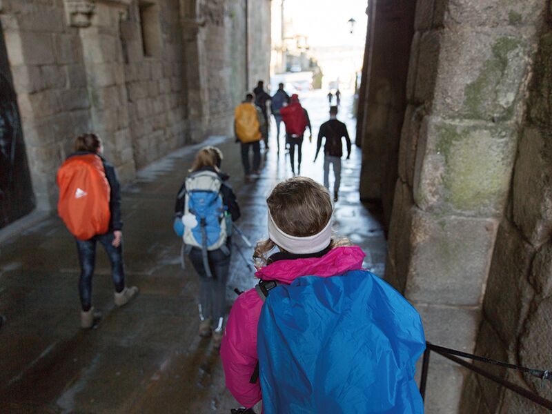 Eine Gruppe von Wandernden mit großen Rucksäcken läuft durch einen steinernen Torbogen in einer alten Stadtgasse.