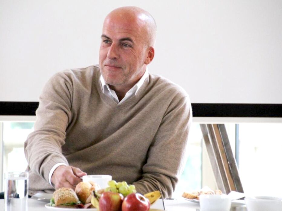 Ein Mann sitzt an einem Tisch mit Obst und Brötchen und schaut nachdenklich zur Seite.