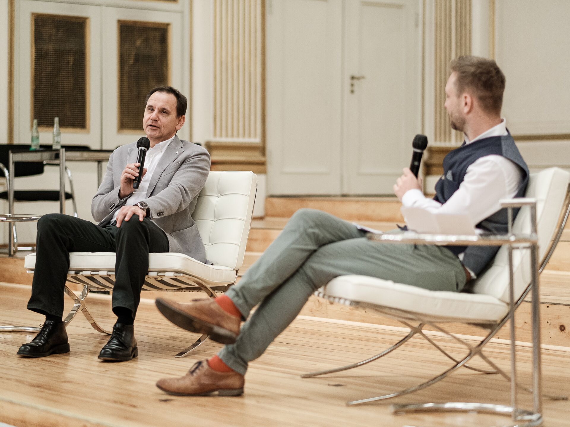Zwei Männer sitzen in weißen Sesseln auf einer Bühne in einem eleganten Saal und sprechen mit Mikrofonen miteinander. Der Mann links trägt ein graues Sakko, der Mann rechts ein weißes Hemd mit grauer Weste.