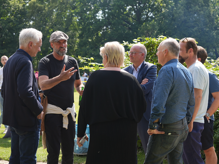 Eine Gruppe von Menschen steht draußen im Grünen beisammen und unterhält sich. Ein Mann mit Mütze und schwarzem T-Shirt gestikuliert lebhaft, während die anderen aufmerksam zuhören.