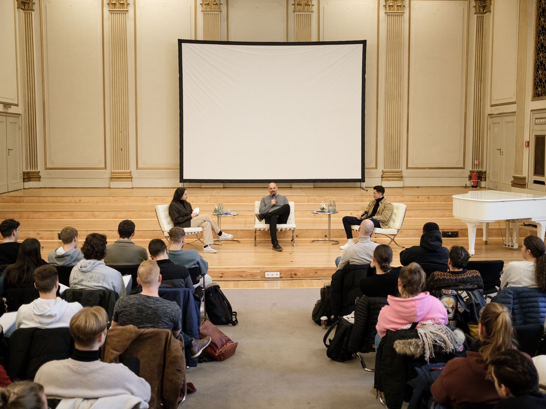 In einem großen Saal diskutieren drei Personen auf einer Bühne vor einer Leinwand, während zahlreiche Zuhörer im Publikum aufmerksam folgen.