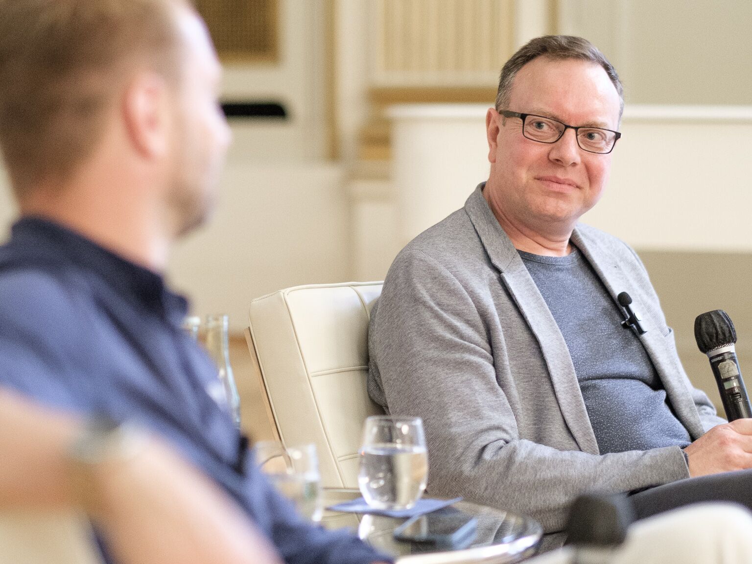 Mann mit Brille und Mikrofon sitzt auf einem Podium und blickt zu einem Gesprächspartner.