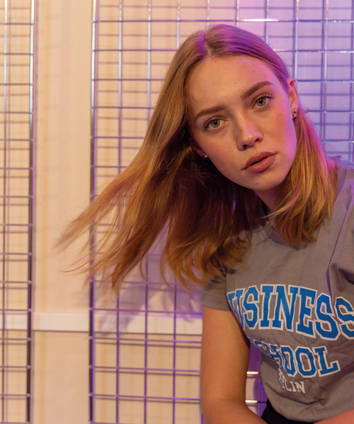 Eine junge Frau mit offenem Haar trägt ein graues T-Shirt mit der blauen Aufschrift „Business School Berlin“ und sitzt vor einem Metallgitter-Hintergrund, der in lila Licht getaucht ist.