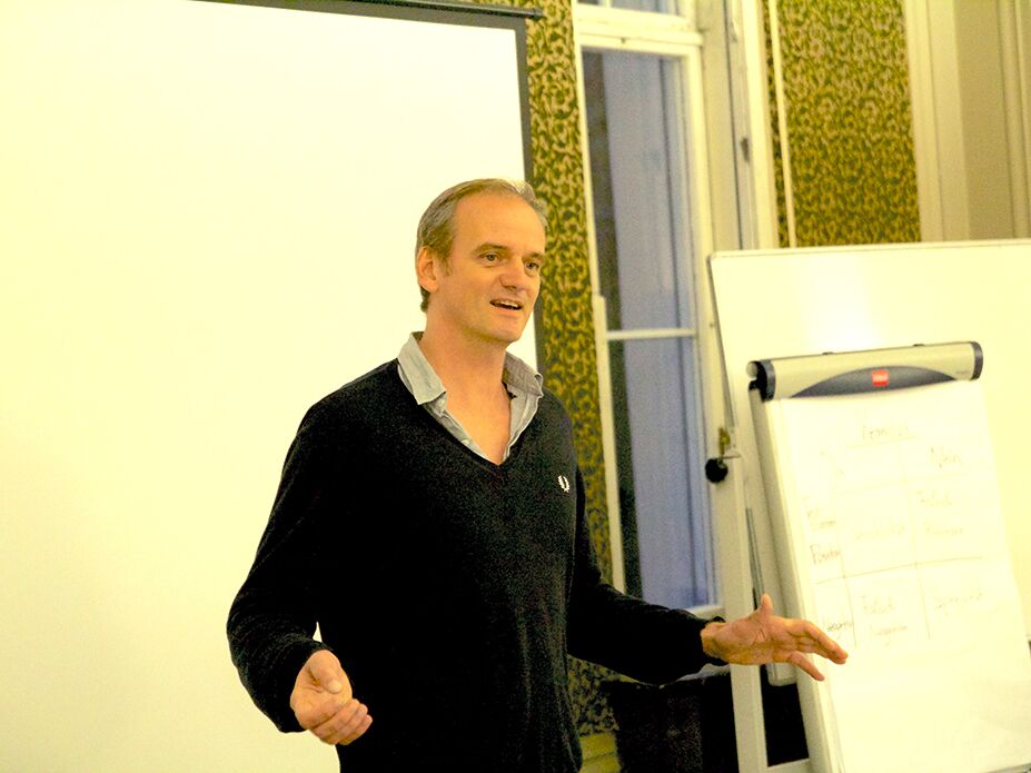 Ein Mann hält einen Vortrag in einem Seminarraum, neben ihm eine Leinwand und ein Flipchart mit Notizen.